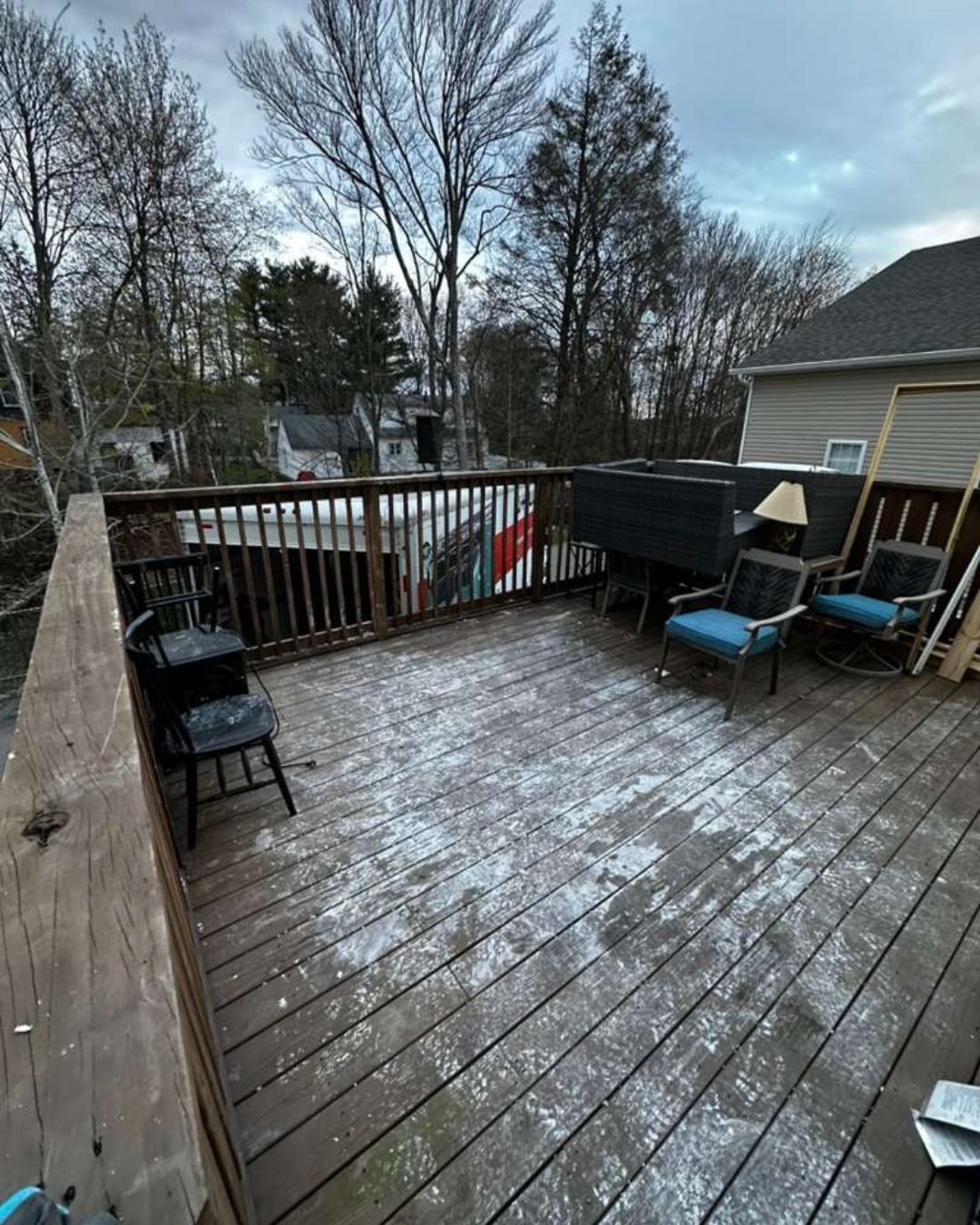 A wet wooden deck cluttered with various pieces of outdoor furniture, including black chairs and blue cushioned patio seats