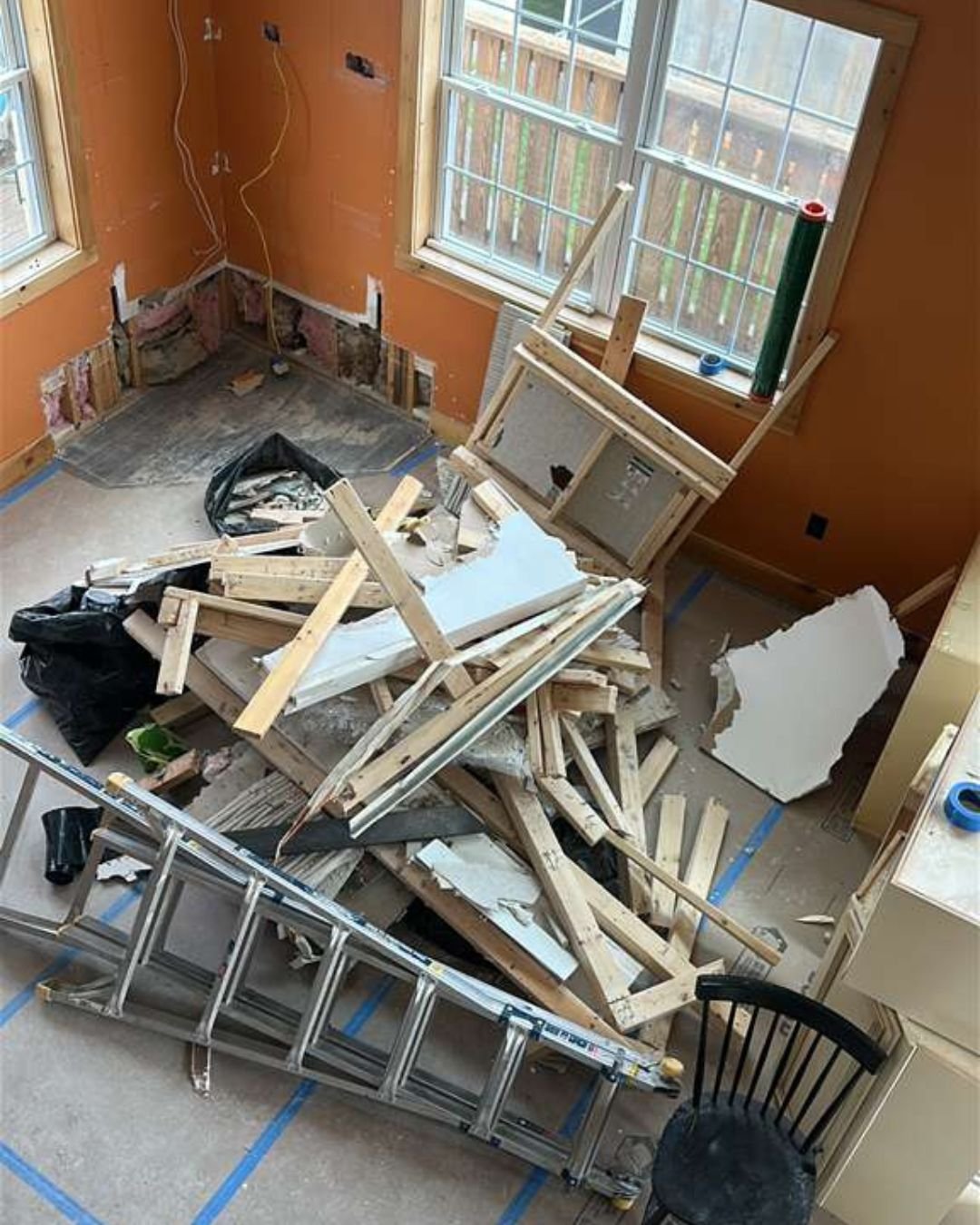 High-angle shot of a room during a renovation project, filled with scattered lumber, drywall scraps, a metal ladder, and a black chair on top of floor protection
