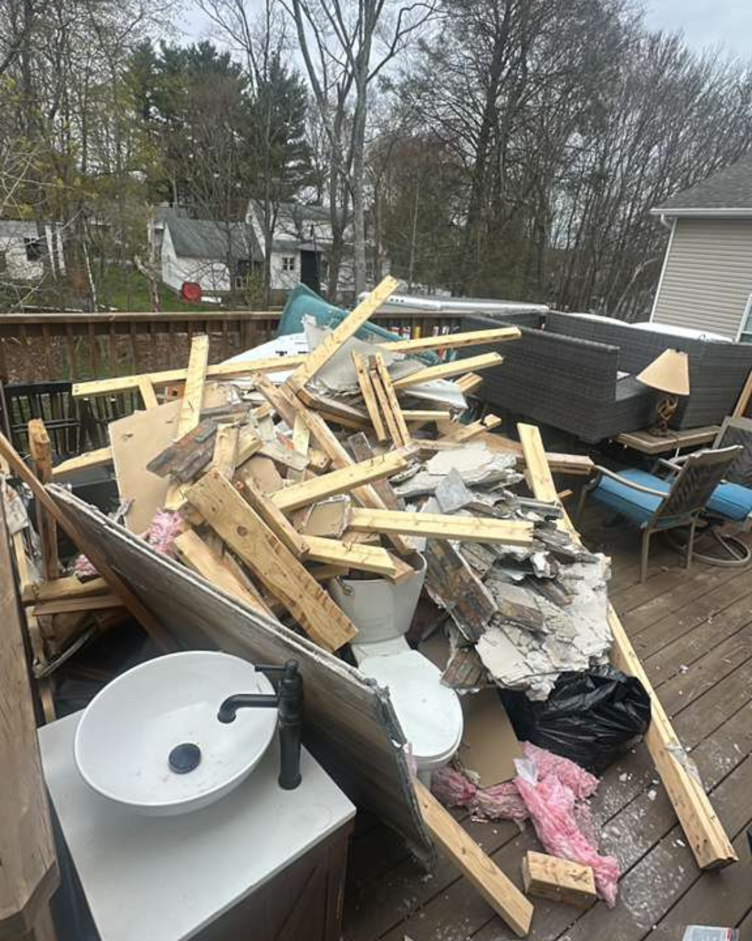 Dramatic piles of wood studs, broken drywall, pink insulation, an old toilet, and a sink vanity scattered across a residential deck during a major renovation