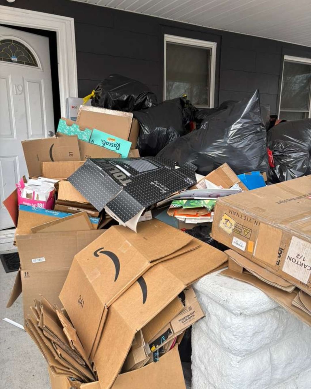 Large stack of flattened and upright cardboard shipping boxes from Amazon and other brands piled on a residential porch.